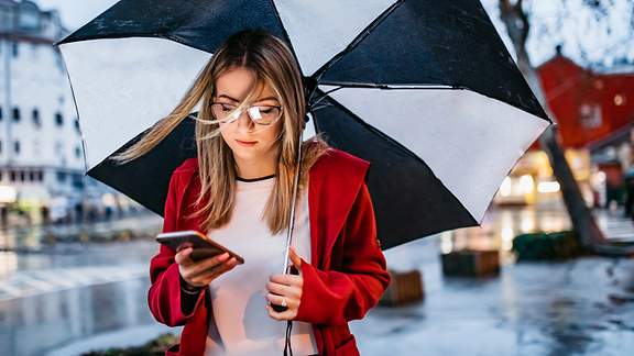 Junge Frau mit Regenschirm blickt auf ihr Handy