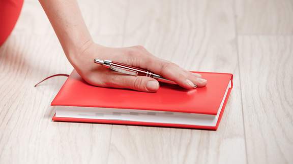 Eine weibliche Hand mit Kugelschreiber liegt auf einem roten Schreibbuch