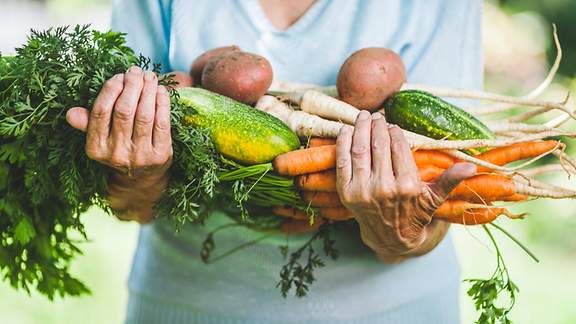 Eine ältere Frau trägt beide Hände voll mit Gemüse vor dem Körper