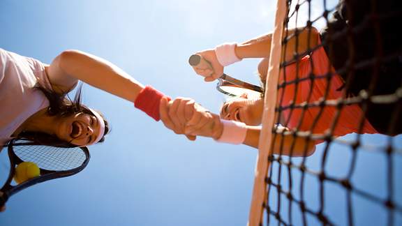 Untersicht auf zwei Tennisspielerinnen, die sich lachend am Netz die Hand geben
