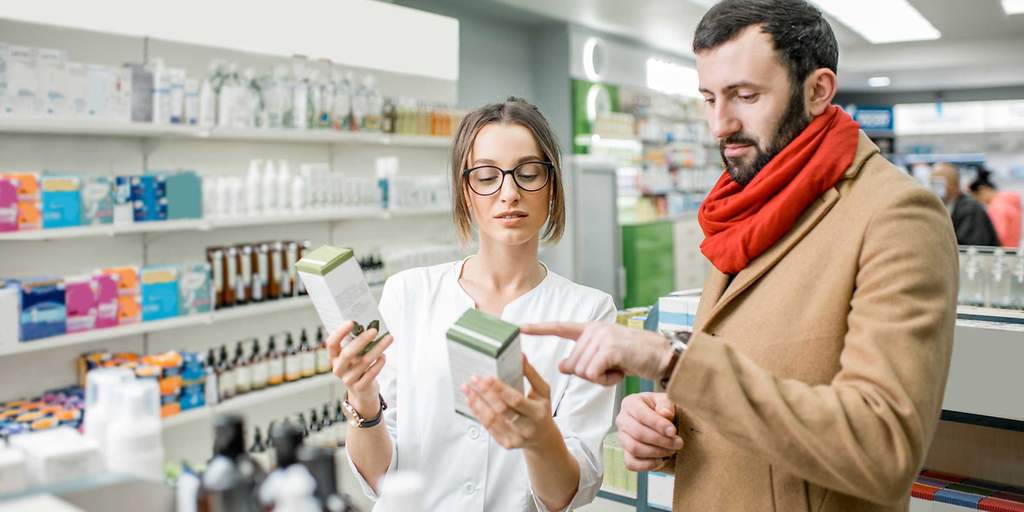 Junge Apothekerin mit Brille berät Mann mit Bart und rotem Schal