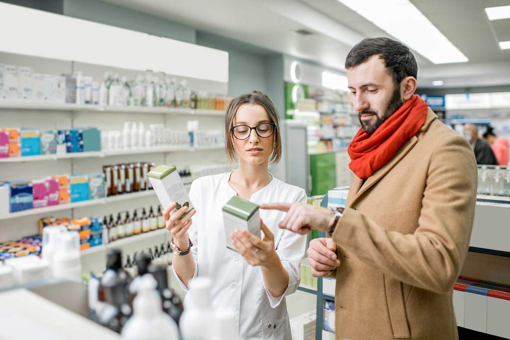 Junge Apothekerin mit Brille berät Mann mit Bart und rotem Schal