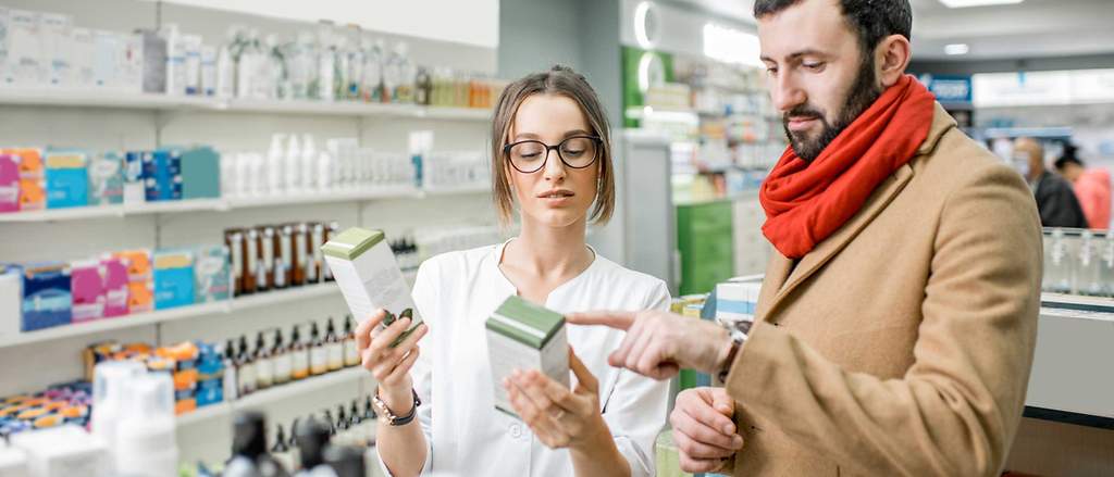 Junge Apothekerin mit Brille berät Mann mit Bart und rotem Schal